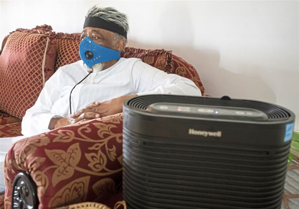 An older man sits on a couch wearing a respirator next to a Honeywell portable air cleaner
