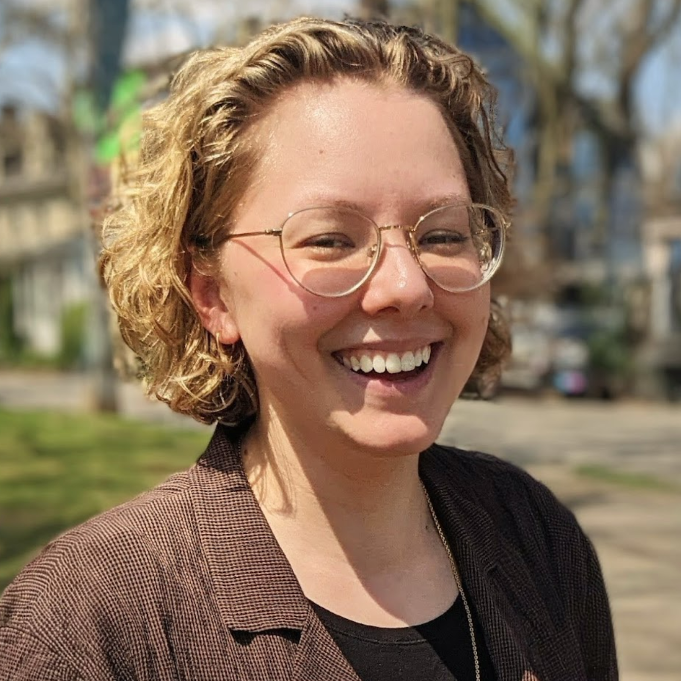 Photo of Elly Helgen, wearing a brown shirt and glasses. She is smiling broadly at the camera.