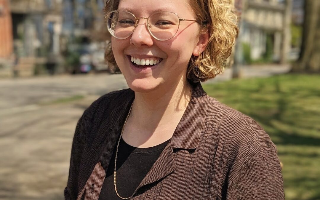 Portrait of Elly Helgen, wearing a brown shirt and glasses, smiling and laughing in an outdoor setting.