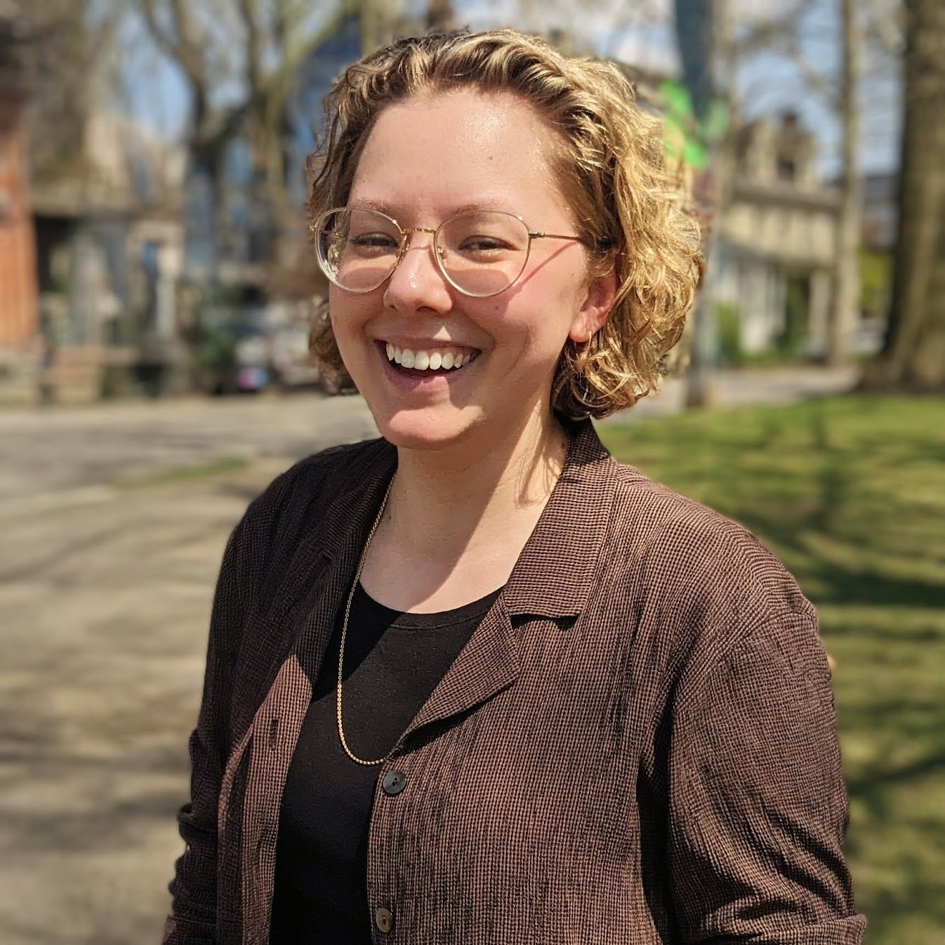 Portrait of Elly Helgen, wearing a brown shirt and glasses, smiling and laughing in an outdoor setting.