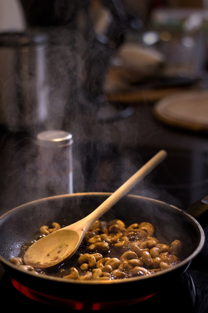 A pan of cashews being roasted on a stovetop. A wooden spoon rests in the pan and smoke rises into the air.