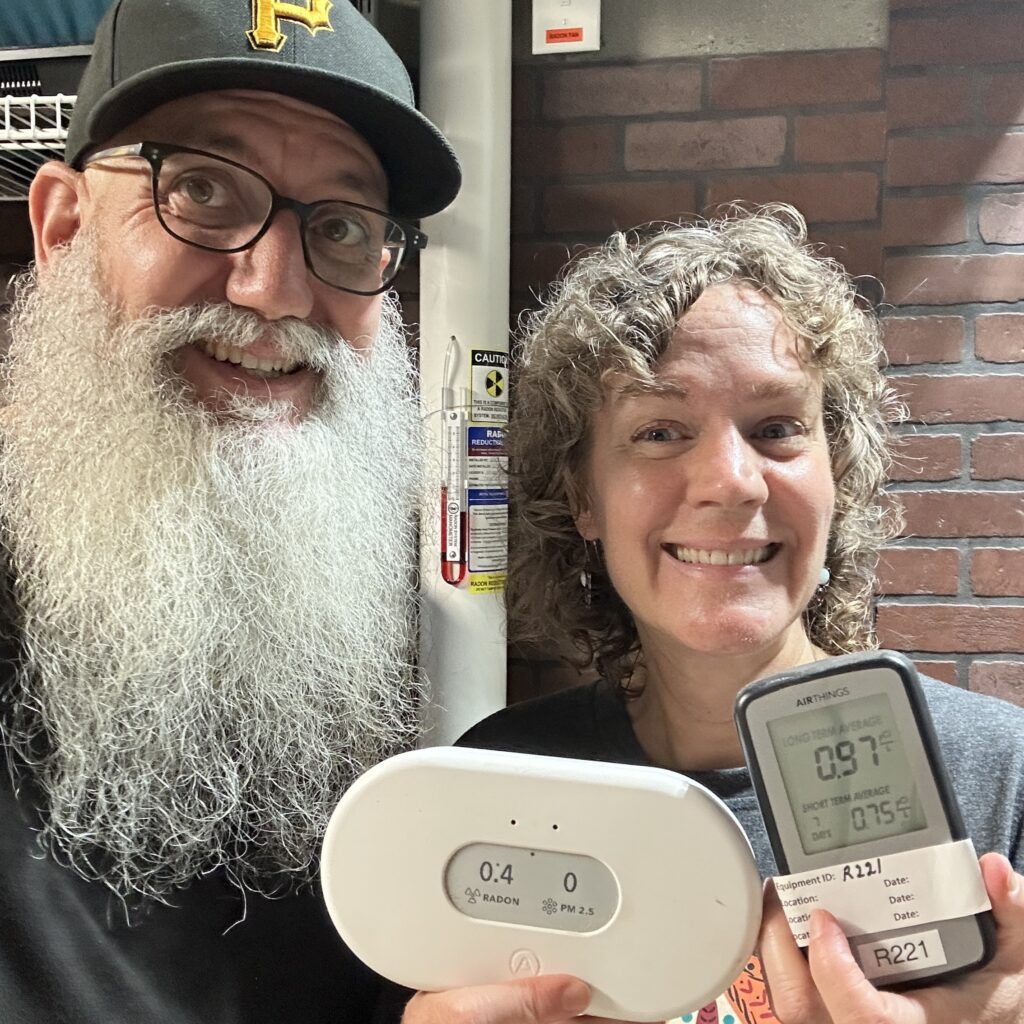 Photo of Karen and Billy in their basement, smiling and holding up air quality monitors